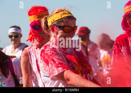 Kaukasische Frau oder eine Frau mit Sonnenbrille und ein gelbes Stirnband rot Pulver Farbe bedeckt Schütteln eine Wolke von Pulver über jemand an einem Sommer Farbe Fun Run Ereignis Stockfoto