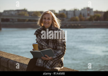 Ältere Frau genießt Buch lesen und Kaffee trinken, während Sie durch den Fluss sitzen. Stockfoto