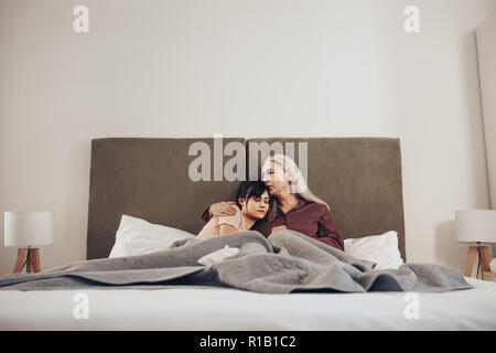 Ältere Frau, die ihre Tochter in den Arm und Küssen mit dem Kopf. Mutter und Tochter auf dem Bett sitzen zusammen. Stockfoto