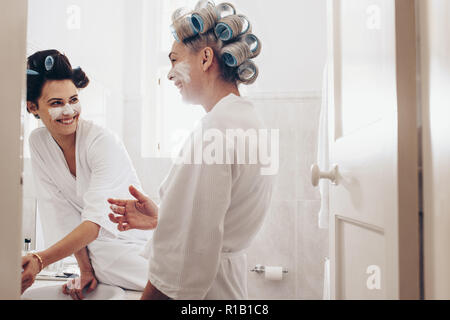 Glückliche Mutter und Tochter stehen im Badezimmer mit Bademänteln mit Curling rollen auf den Kopf. Frauen Anwendung Gesichtscreme stehend im Bad bei h Stockfoto