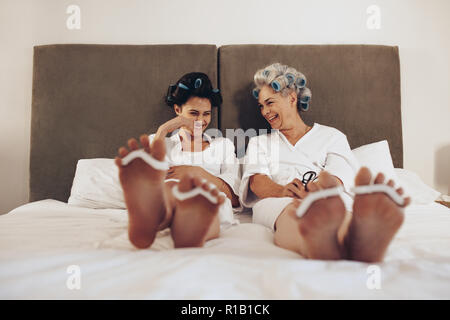Glückliche Mutter und Tochter sitzen am Bett mit Curling Rollen fixiert auf das Haar. Zwei Frauen sitzen auf dem Bett entspannen beim Pflegen Haar und Füße. Stockfoto