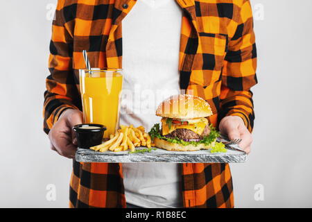 Student in einem karierten orange Hemd auf einem weißen Hintergrund ist ein Holzbrett mit einem frischen Burger, ein Glas Orangensaft und Bratkartoffeln. Das Konzept der ungesunden Ernährung Stockfoto