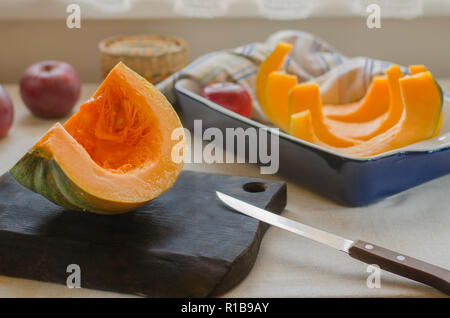 Eine orange-grün Kürbis liegt auf einem braunen Holz Schneidebrett und in einem blauen Keramik Auflaufform Stockfoto