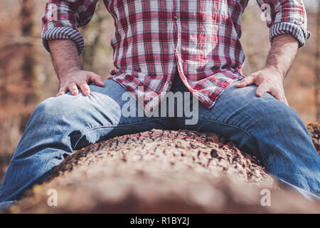 Starke Mann im karierten Hemd und blauer Jeans sitzt auf einem Baumstamm, Beine gespreizt Stockfoto