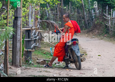 Don Det, Laos - April 23, 2018: Junge buddhistische Mönch sitzt auf einem Roller und über sein Handy in einem abgelegenen Dorf im südlichen Laos Stockfoto