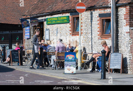 Junge Kellnerin, die im Café Flintstones Tearooms in Emsworth, Hampshire, England, Großbritannien, Essen an Gäste bringt, die draußen sitzen. Stockfoto