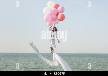 Junge Frau in einem weißen Anzug schwebt in der Luft in Luftballons. Stockfoto