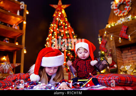 Bezauberndes Mädchen in Santa Hut auf dem Boden liegen und schreibt Schreiben, Zeichnen mit Bleistift und ihr kleiner Bruder, die mit Ihr stört, spielt mit Bleistifte in warmen Raum mit Girlanden, Leuchten Stockfoto