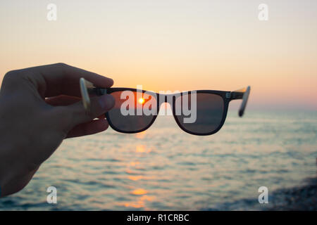 Sonnenbrille in der Hand des Menschen auf den Sonnenuntergang. Die Sonne scheint durch die Brille Stockfoto