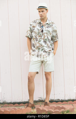Ein stattlicher Mann, männliches Modell im Sommer Kleidung im Freien stehend gegen eine einfache hölzerne Wand posieren. Er trägt einen weißen Hut. Stockfoto
