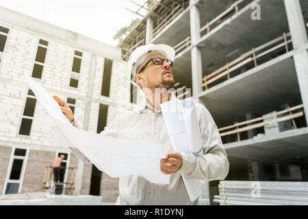 Bauingenieur in einem weißen hardhat Inspektion Blueprints auf einer Baustelle. Entwicklung und Konstruktion Industrie Konzept Stockfoto