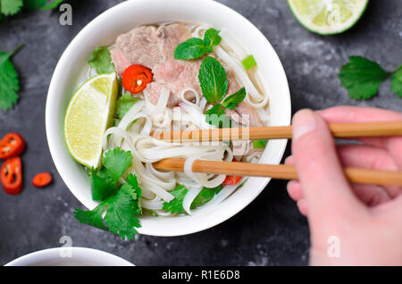 Traditionelle vietnamesische Suppe Pho Bo mit Reis Nudeln, Rindfleisch und Kräutern auf dunklem Hintergrund, asiatische Küche, Chopstick in weiblicher Hand Stockfoto