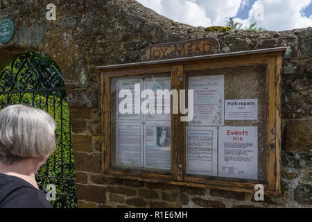 Ältere Frau liest das Information Board für Freude Mead Gärten im Dorf Farthingstone, Northamptonshire, Großbritannien Stockfoto