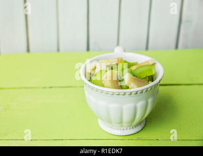 Frische reife Rhabarber (Rheum rhabarbarum), geschält und in Stücke in einem weißen vintage Schale schneiden. Für das Kochen vorbereitet. Grün und weiß Holz- Hintergrund. Stockfoto