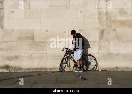 Einsamer Mann, Mann, Erwachsener, mit Fahrrad gegen eine ebene Wand Hintergrund an einem sonnigen Tag. Mit smartphone, Mobiltelefon, Kopf nach unten, texting, während Taube durch Stockfoto