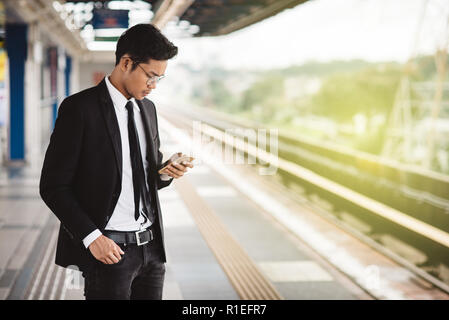 Jungen asiatischen hipster Geschäftsmann mit Smartphone warten am Bahnsteig. Konzept der drahtlosen Technologie, den öffentlichen Verkehrsmitteln und Business Travel Stockfoto