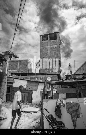 Schwarzer Mann zu Fuß durch den Slum von Rio de Janeiro gebaut von einfachen Hütten mit schwarzem Rauch in den Himmel, die bei der Verbrennung von Müll Stockfoto