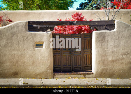 Eine bezaubernde Aussicht auf eine Adobe Wand- und Vintage Holztüren mit roten Blättern auf Bäume im Herbst Innenhof im historischen Viertel von Santa Fe, NM Stockfoto