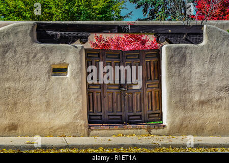 Eine bezaubernde Aussicht auf eine Adobe Wand- und Vintage Holztüren mit roten Blättern auf Bäume im Herbst Innenhof im historischen Viertel von Santa Fe, NM Stockfoto