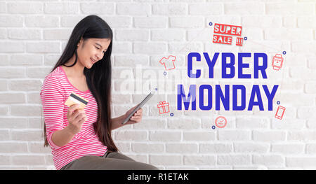 Asiatische Frau in ungezwungener Tuch halten und mit Kreditkarte einkaufen in der Cyber Monday im Verkauf mit Tablet am weißen Backsteinmauer online. Digitale Technologie l Stockfoto