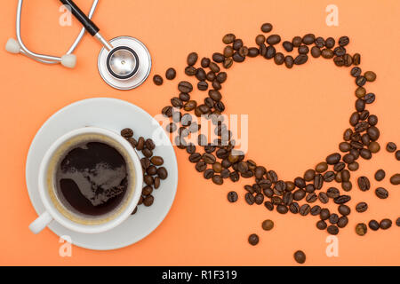 Weißes Porzellan Tasse Kaffee auf Untertasse mit Kaffeebohnen in der Form eines Herzens und phonendoscope auf Pfirsich farbige Hintergründe gefaltet. Ansicht von oben Stockfoto