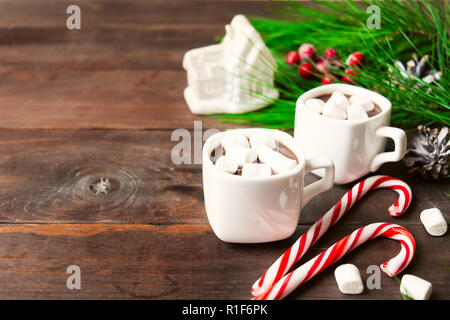 Zwei heiße Getränke mit Marshmallow in Cups mit Spielzeug Haus und zwei Zuckerstangen, Weihnachten Getränke auf Holz- Hintergrund mit Kopie Raum Stockfoto