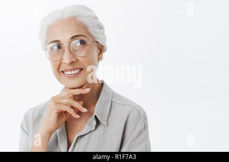 Smart kreative und erfreut, charmante Granny mit weißen Haaren in Schaugläser neugierig lächelnd die Hand oben Kinn Blick auf die rechte obere Ecke, beim Denken der Entscheidung beachten Stockfoto