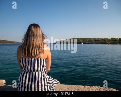 Junge Frau mit schönem Haar sitzt auf Stein Dock in der Nähe des Meeres auf der Insel Vis in Kroatien Stockfoto