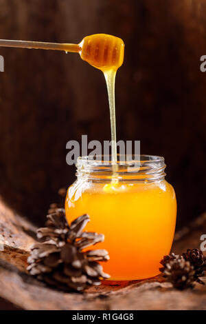 Die duftenden Honig fließt von einem Stick in ein Glas vor dem Hintergrund der Rinde und Zapfen. Close-up. Stockfoto