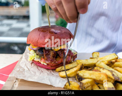 Riesige saftige Burger mit verschiedenen Toppings und schmackhaftes Fleisch serviert mit großen hausgemachten Pommes frites auf einer Holzplatte. Männliche Hand, gebratene Kartoffeln mit Gabel. Stockfoto