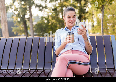 Junge Frau liegt im Herbst Park auf der Bank nach einem Spaziergang und hört die Musik auf das Telefon an einem sonnigen Tag Stockfoto