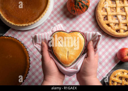 Frau mit einem Raspberry pie, in einer herzförmigen Fach, über einen Küchentisch voll von traditionellen fallen, Torten, Kuchen, Apfel und Kürbis Kuchen. Stockfoto