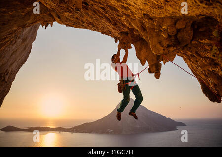 Männliche Kletterer hängen auf einer Klippe mit einer Hand bei Sonnenuntergang. Insel Kalymnos, Griechenland. Stockfoto
