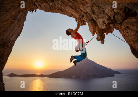 Männliche Kletterer hängen mit einer Hand auf die anspruchsvolle Strecke auf einer Klippe bei Sonnenuntergang Stockfoto