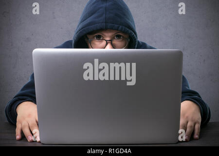 Hacker in Gläsern mit großen Augen auf einem konkreten Hintergrund sitzt an einem Laptop und mit Blick auf die Kamera Stockfoto