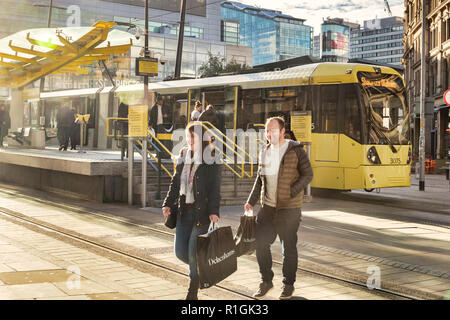 2. November 2018: Manchester, Großbritannien - Junges Paar lächelnd, mit Debenhams Einkaufstaschen, in Exchange Square, mit einem Metrolink tram hinter sich. Viel Flair. Stockfoto