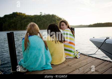 Mädchen sitzen auf einem Steg mit ihren zwei Freundinnen tragen Handtücher. Stockfoto