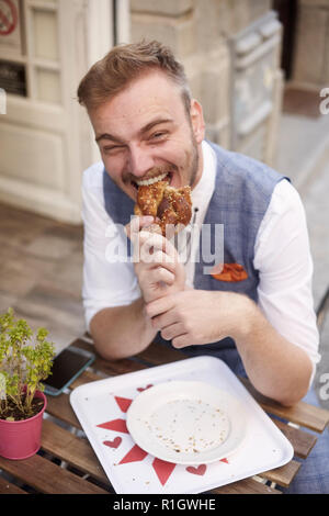 Eine junge lächelnde Mann, 20-29 Jahre alt, trug hipster Anzug, Standortwahl auf Straße, brezel Essen vor der Bäckerei im Freien. Seltsam, lustiges Gesicht ein Stockfoto