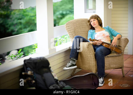 Junges Mädchen sitzt auf einem Korbstuhl auf einer Veranda, während eine Geige. Stockfoto