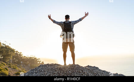 Ansicht der Rückseite des jungen Mann mit Rucksack steht auf der Spitze des Berges mit ausgebreiteten Armen öffnen. Man genießt die Aussicht von der Spitze des Hügels. Stockfoto