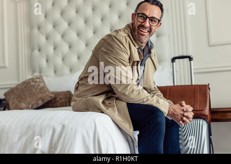 Erfolgreicher Geschäftsmann mit Gepäck im Hotel Zimmer Bett sitzen und Wegsehen. Reifen Geschäftsmann auf Geschäftsreise. Stockfoto