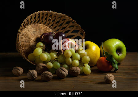 Das Stillleben des umgedrehten Obstkorbes mit den verstreuten Früchten auf der braunen Oberfläche. Schwarzer Hintergrund. Dramatische Beleuchtung. Stockfoto