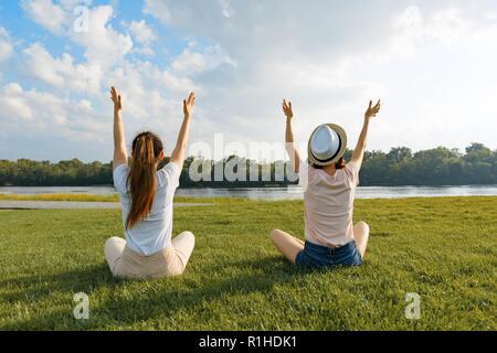 Zwei junge Mädchen im Park in der Nähe des Flusses meditieren, zurück, die Mädchen ihre Hände oben angehoben, goldene Stunde. Stockfoto