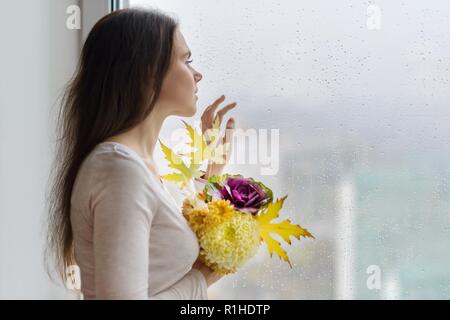 Hallo Herbst. Junge lächelnde Frau mit Herbst Blumenstrauß aus gelben und orangefarbenen Blüten, violett Zierpflanzen Kohl und Ahornblätter. Weibliche in der Nähe der Fenster Stockfoto