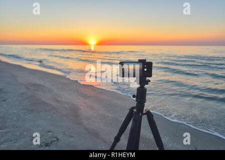 Sonnenaufgang über dem Meer. Feuerball der Sonne über dem Horizont in einem farbenfrohen Orange Sky. Smartphone Kamera auf einem Stativ im Vordergrund. Stockfoto