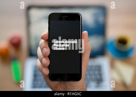 Ein Mann schaut auf seinem iPhone die zeigt die International Business Times logo, während saß an seinem Computer Schreibtisch (nur redaktionelle Nutzung). Stockfoto