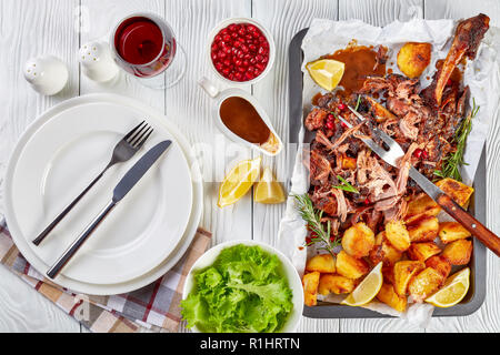 Ansicht von oben langsam Braten ultra-zarte Lammkeule zog die Knochen mit knusprigen Braten Kartoffeln auf ein Backblech auf einem Holztisch mit Glas Stockfoto