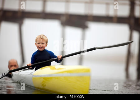 Junge Junge lächelt als er hält ein Ruder und sitzt in einem Kajak als Mann es entlang drückt in den Gewässern Eines nebligen Hafen. Stockfoto