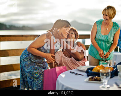 Lächelnd, reife Frau Freund küsst sie auf die Wange in Gruß, als sie für ihre Geburtstagsparty in einem Restaurant am Meer ankommt. Stockfoto