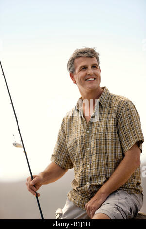 Lächelnd im mittleren Alter Mann sitzt auf einem Felsen mit einer Angelrute. Stockfoto